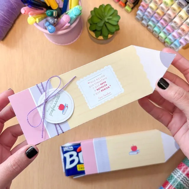 Embalagem em formato de lápis amarelo, rosa e lilás com frase de Cora Coralina e tag Feliz dia dos professores amarrada com cordão lilás.